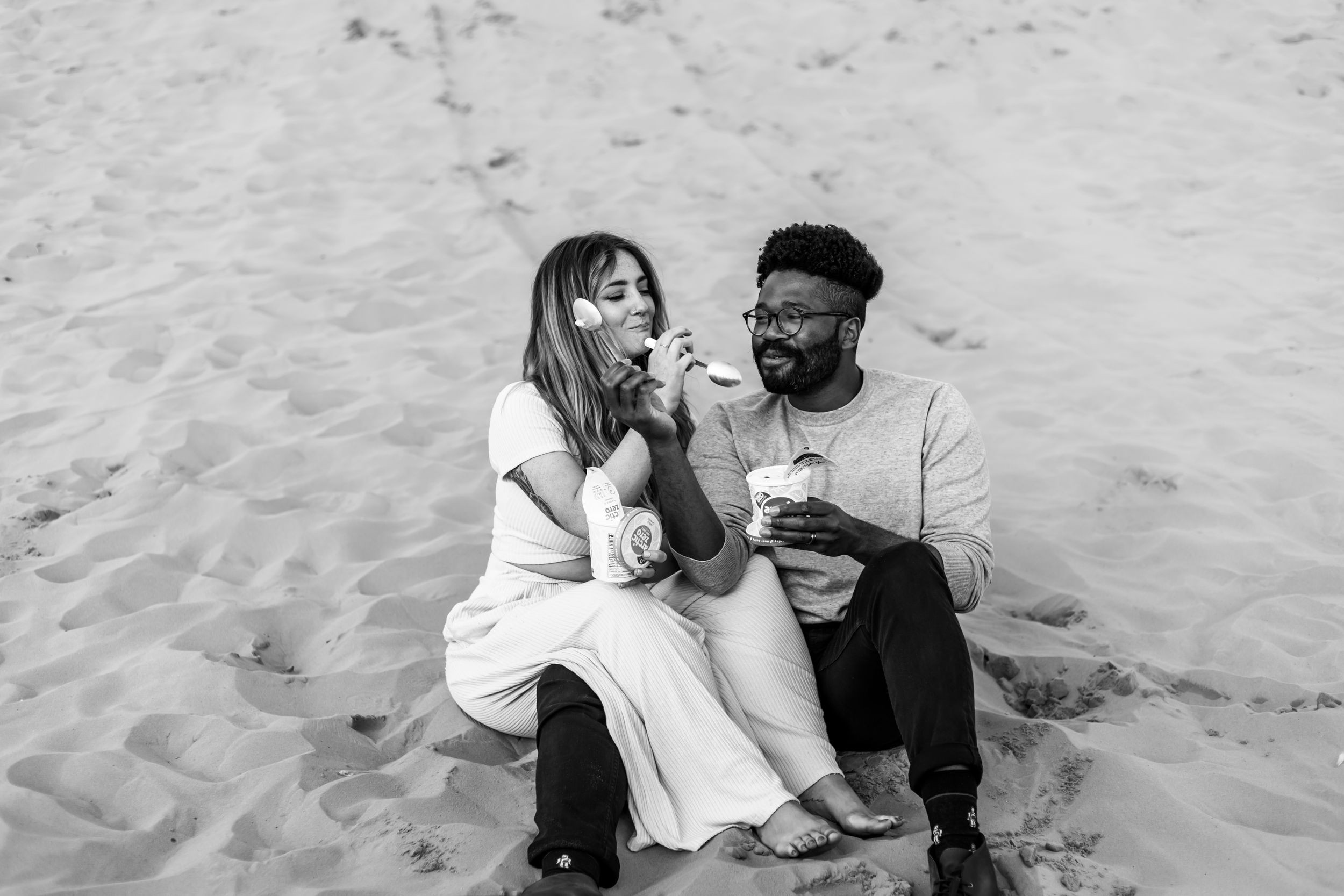 A couple shares a joyful moment while sitting on sand dunes in black and white photography.