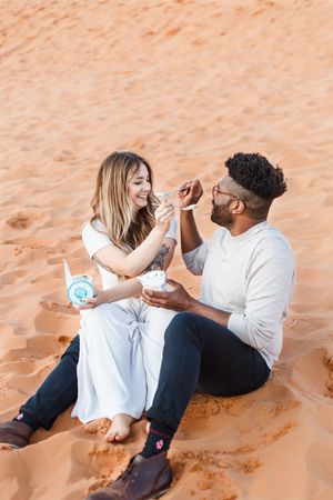 Couple sits playfully in orange sand dunes while sharing a romantic moment in casual white and black outfits.