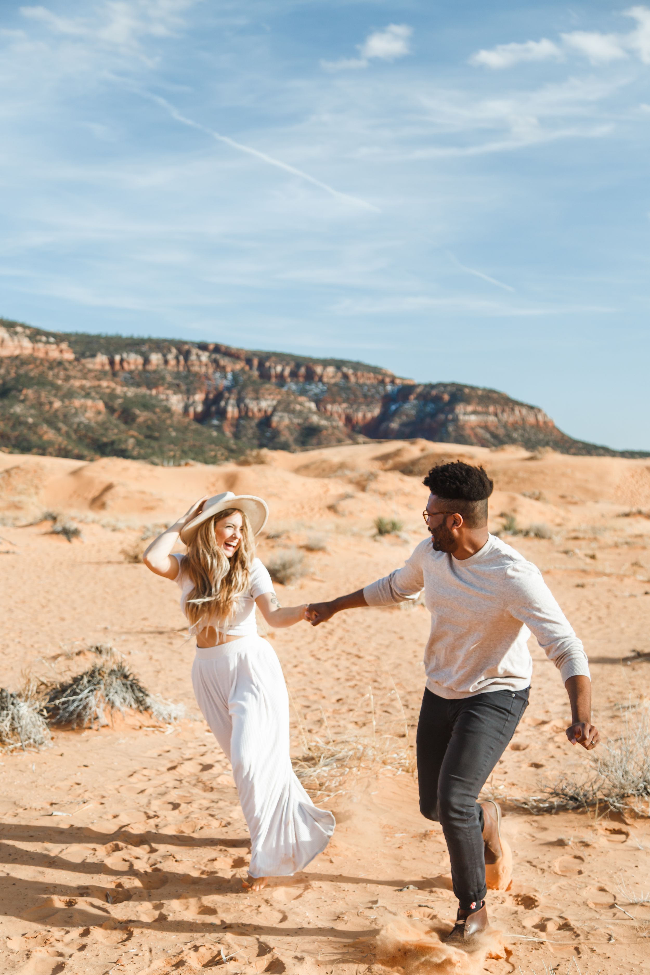 A couple playfully runs and dances together on orange sand dunes with rocky cliffs in the background.