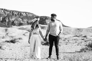 Couple walking hand in hand through scenic desert landscape with mountains in background at sunset.