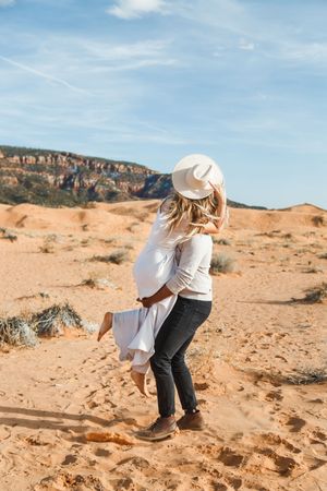 Person in white shirt and hat exploring red rock desert landscape against blue sky and cliffs in background.