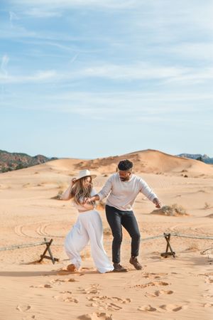 A romantic couple poses playfully on orange sand dunes against a bright blue desert sky.