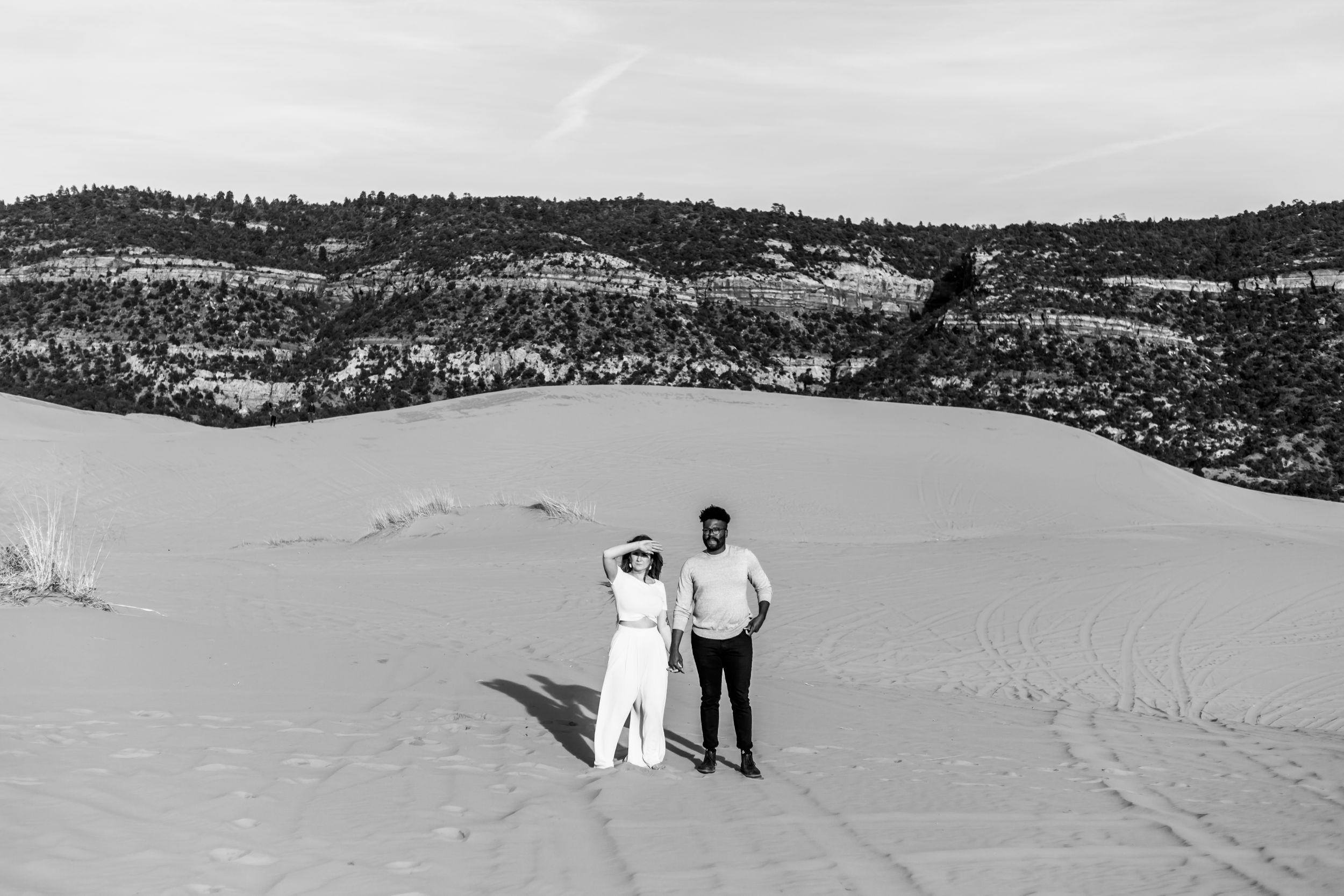 A couple stands together on a scenic sand dune overlooking a vast desert landscape in black and white.