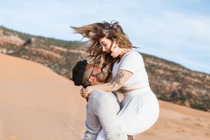 Playful couple embracing and laughing together on sandy dunes with mountain landscape behind them.