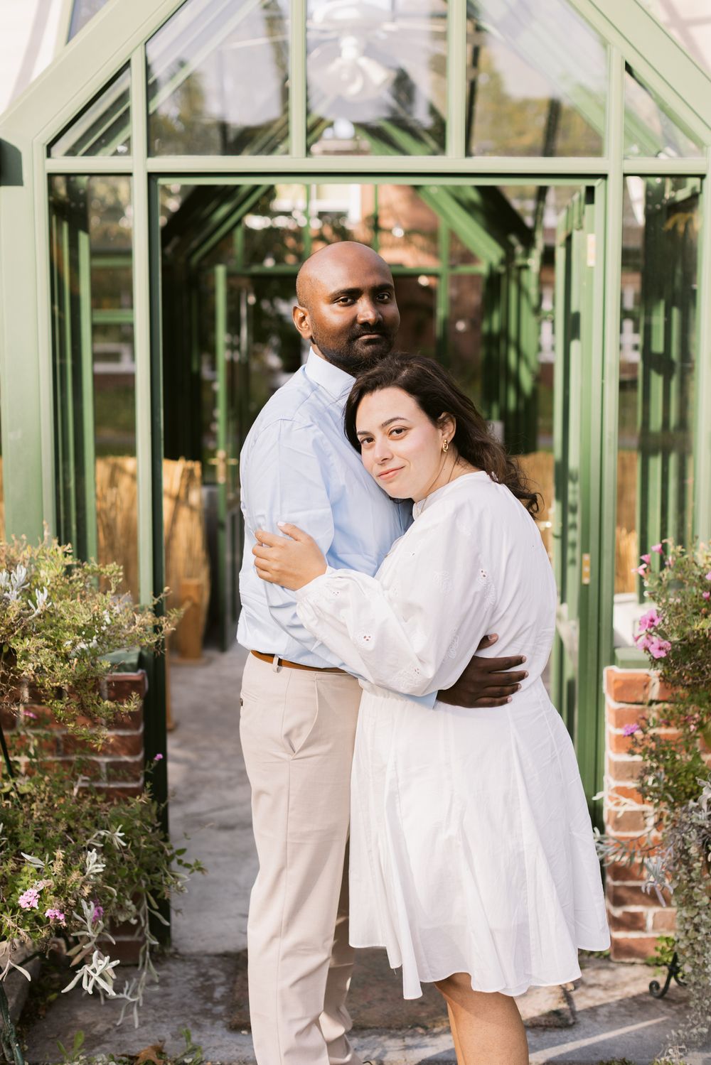Man wears blue shirt embraces woman wearing white dress standing in front of glass greenhouse for couple photos. Harvard couple photographer
