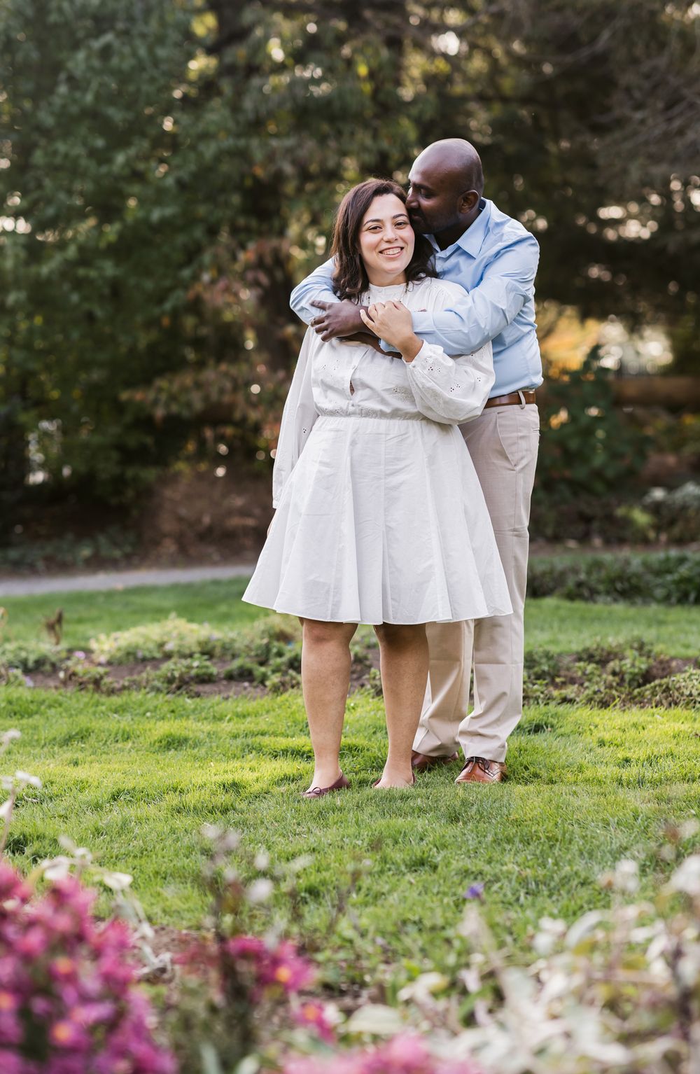 Man in blue shirt embraces woman wearing white dress from behind at Garden at Elm Bank in Wellesley. Newburyport couple photographer