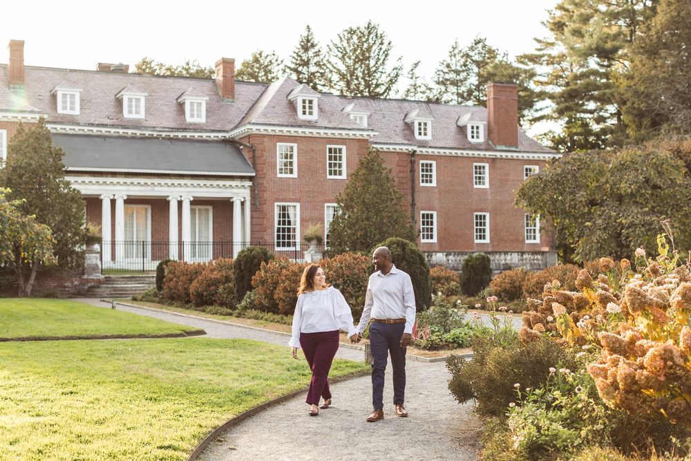 A couple walks on a pebble walkway holding hands in front of large colonial brick building at Garden at Elm Bank in Wellesley, Massachsuetts