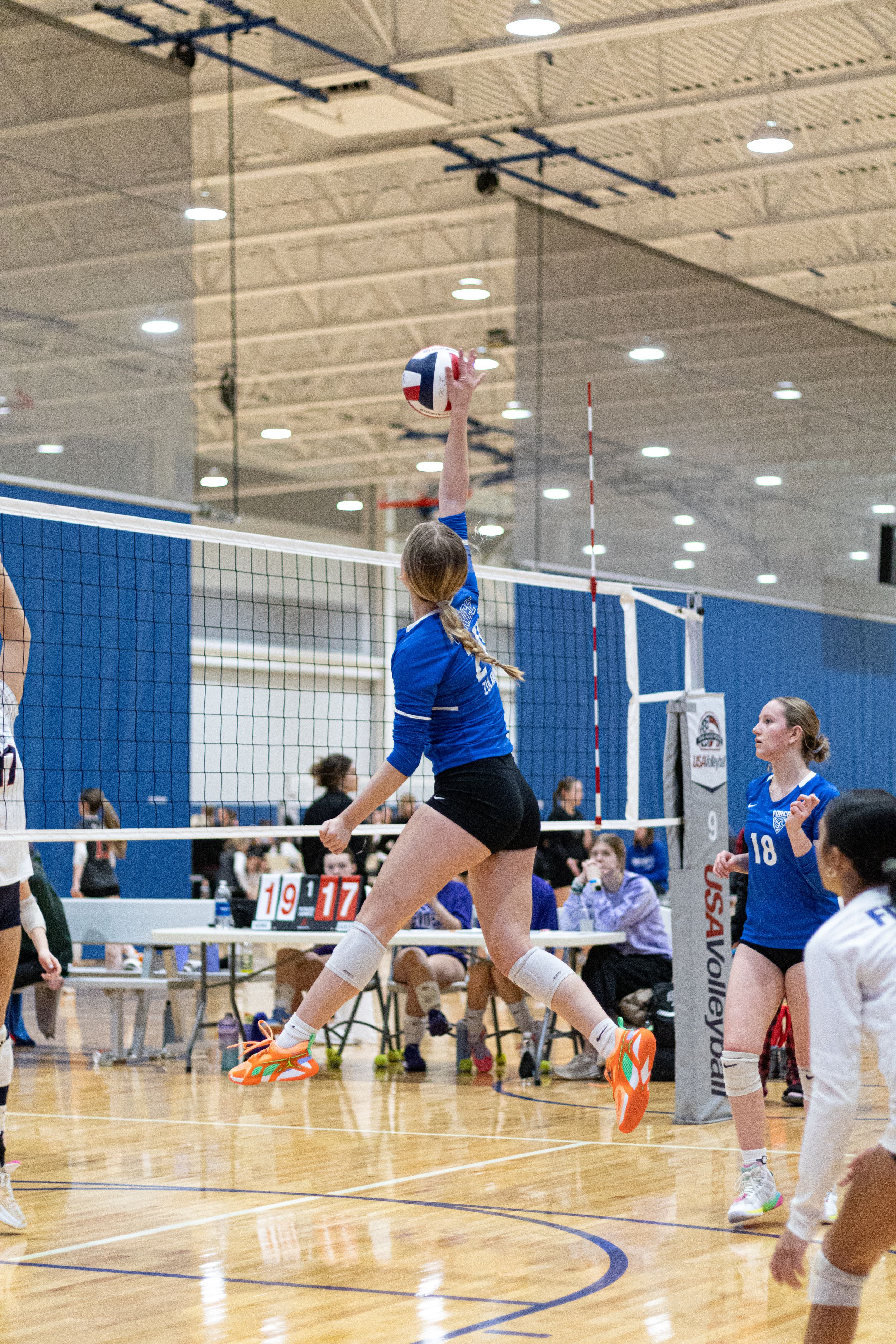 Action shot of a volleyball player in a blue jersey jumping high to spike the ball over the net.
