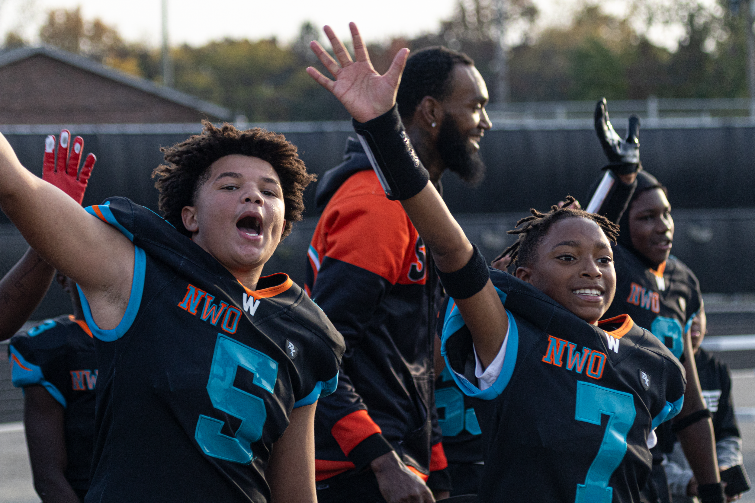 Excited youth football players in black and teal jerseys cheering with arms raised on the field.