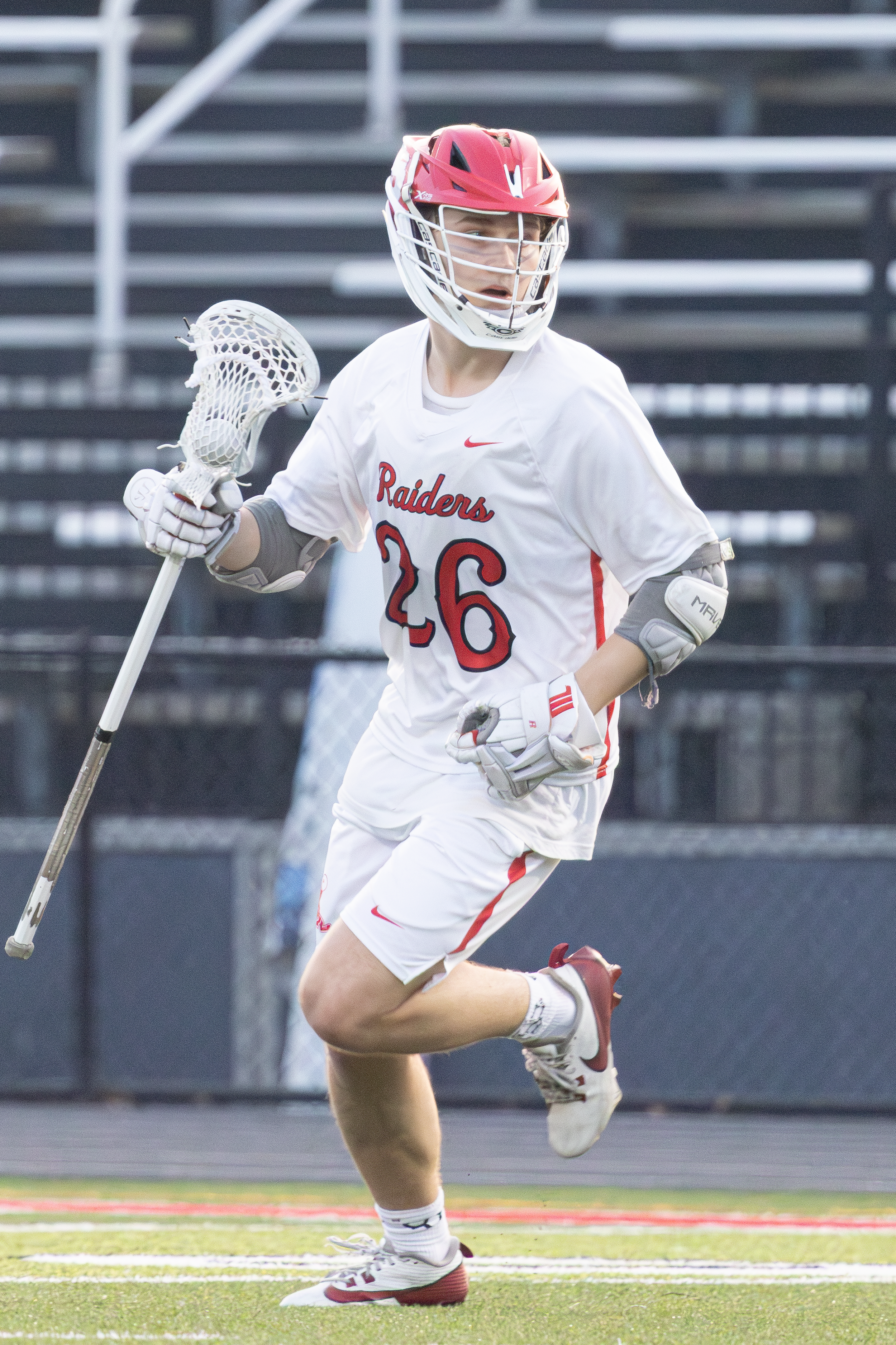 Lacrosse player in a white Raiders jersey and red helmet running across a turf field with his stick in hand.