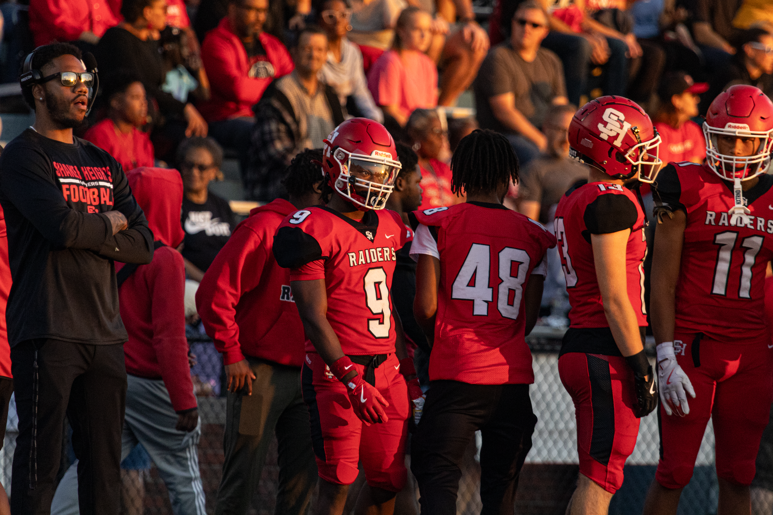 High school football players in red Raiders jerseys standing on the sideline with their coach at sunset.