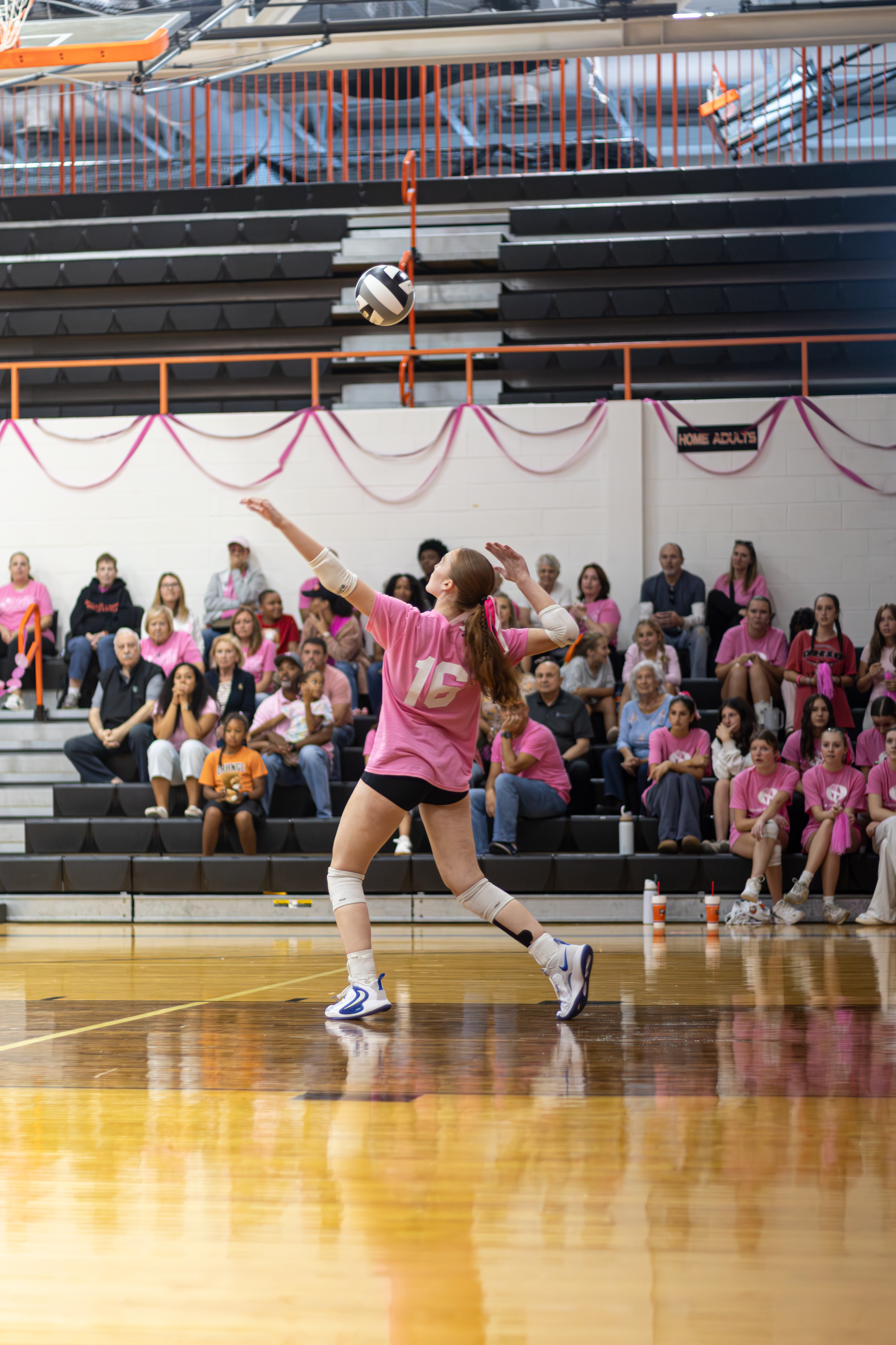 Volleyball player in a pink jersey serving the ball in a gym during a pink-out event.