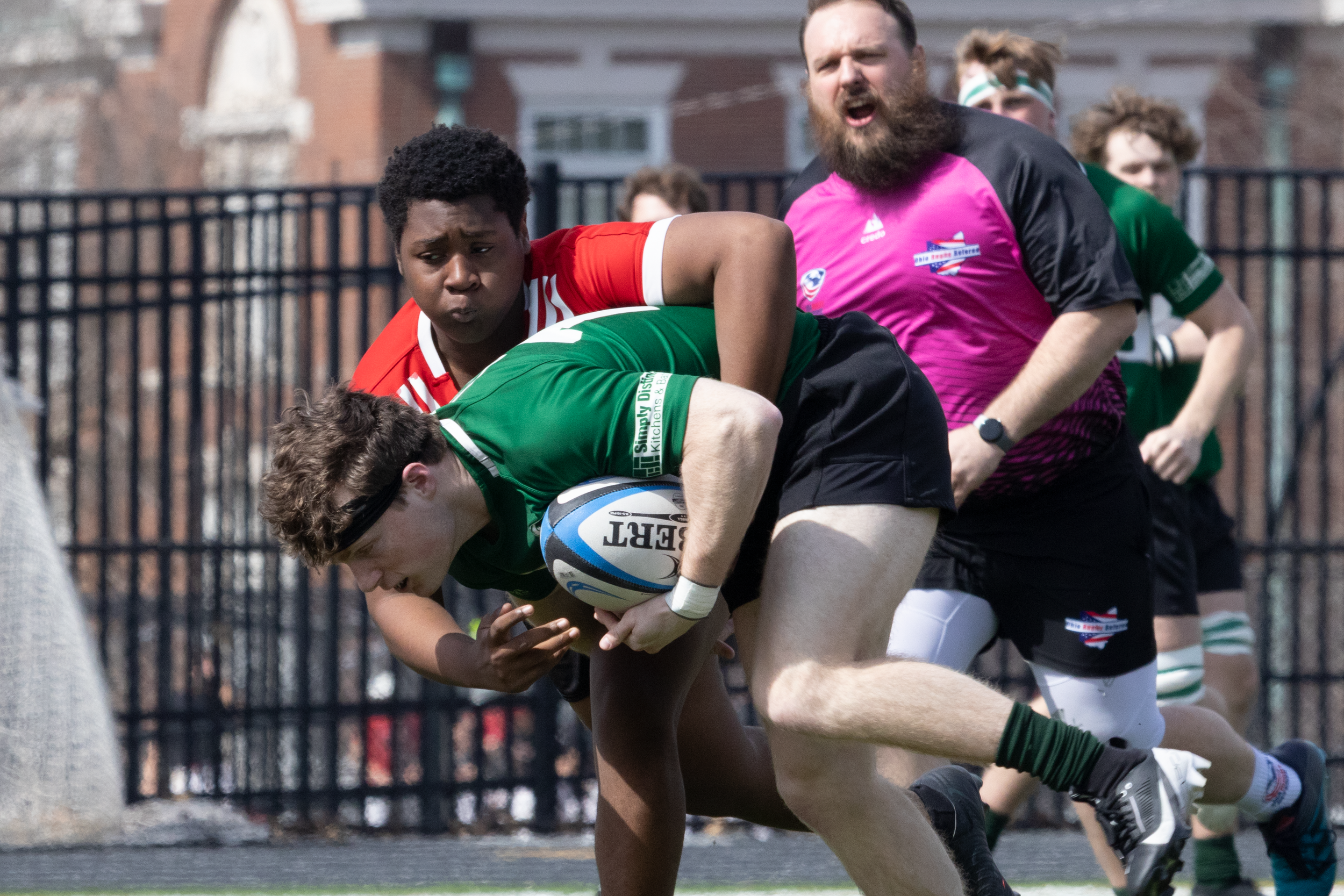Close-up of a rugby tackle with a green jersey player carrying the ball and a referee in pink behind them.