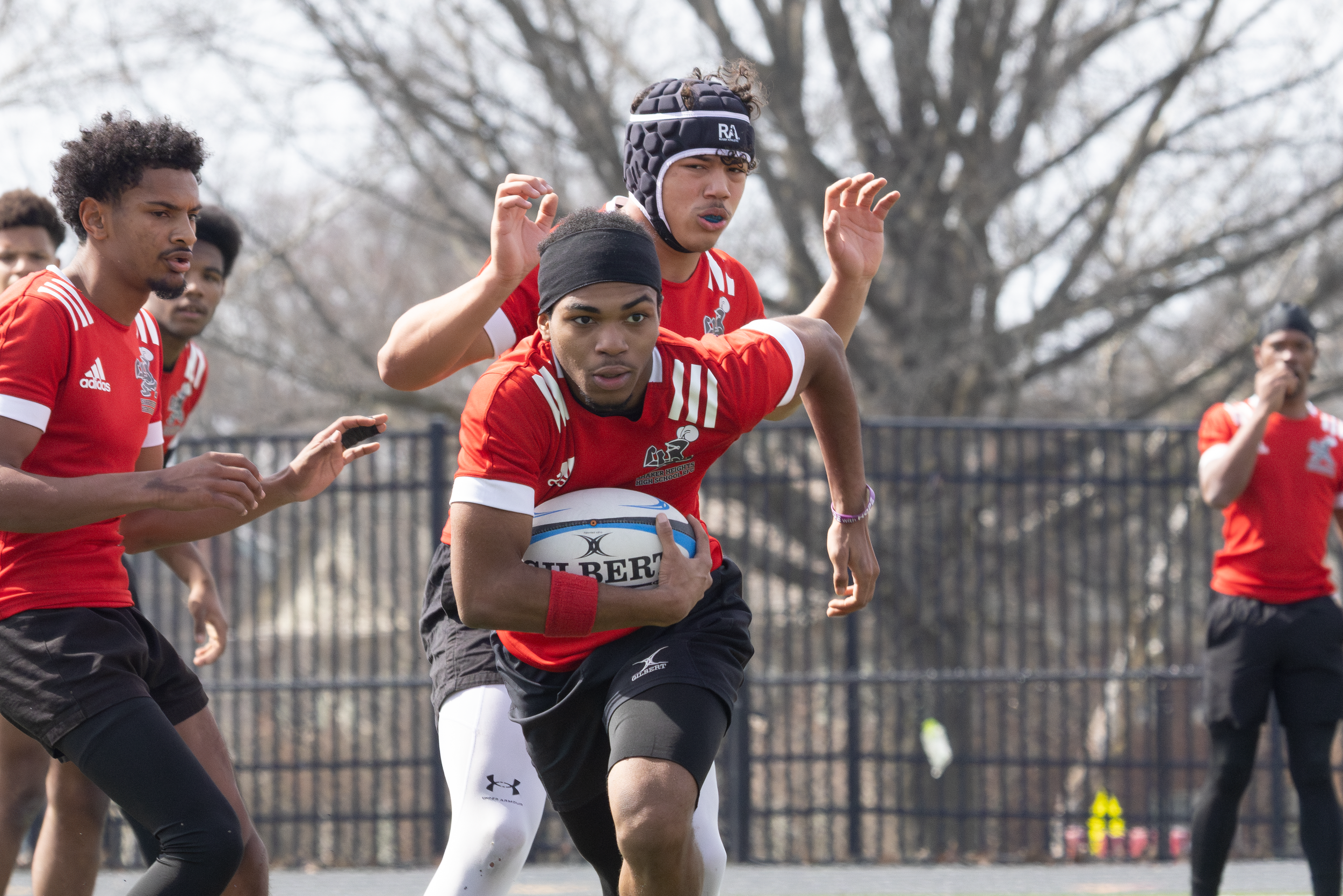 Rugby player in a red jersey sprinting with the ball during an outdoor match.