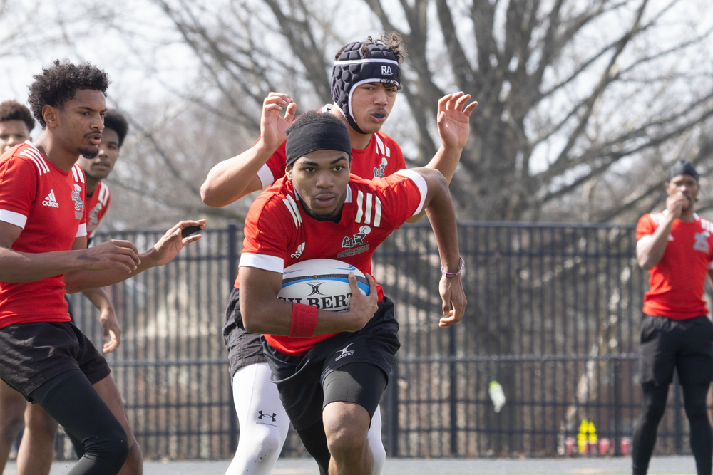 Rugby player in a red jersey sprinting with the ball during an outdoor match.