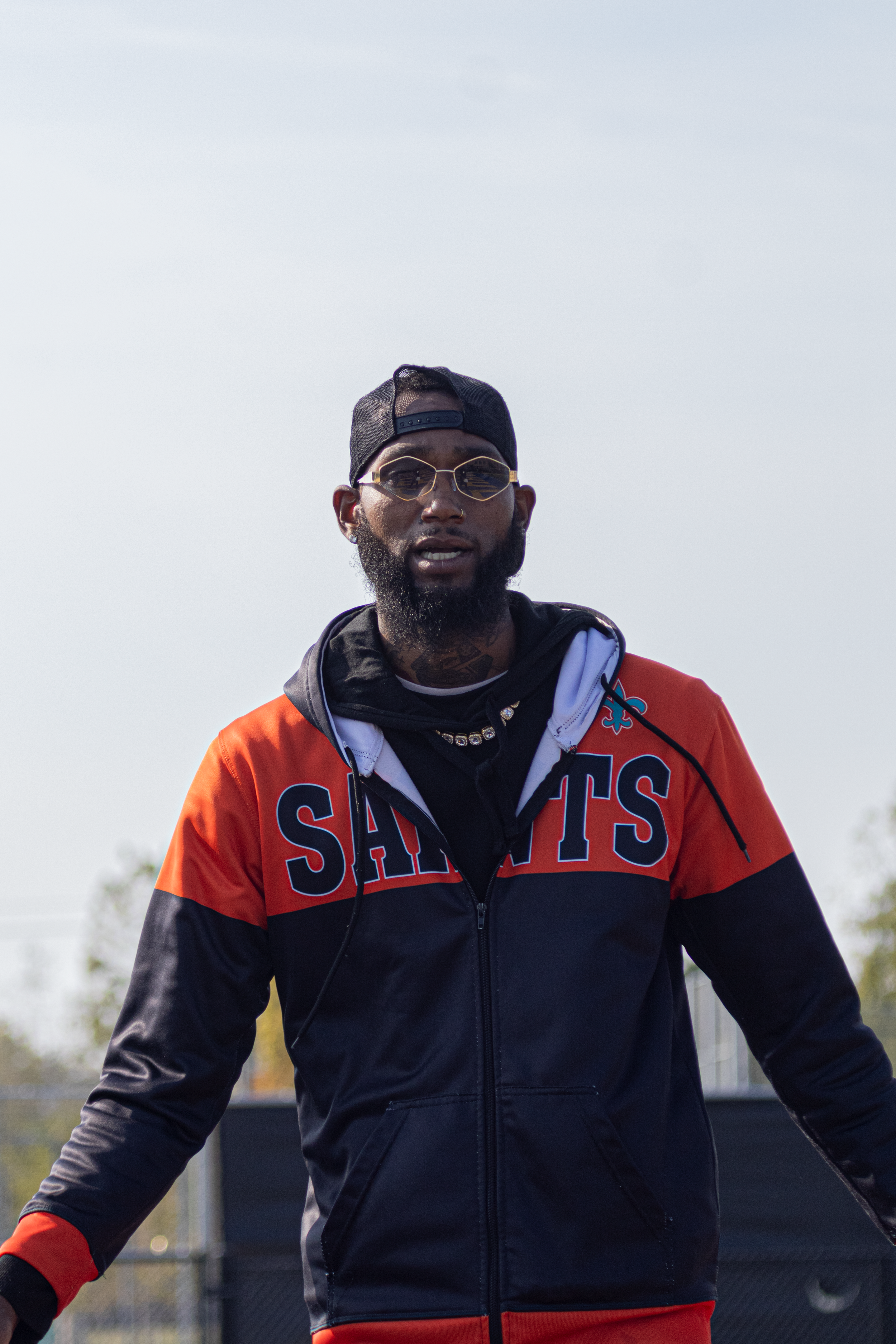Portrait of a man in an orange and black Saints jacket with gold glasses and a black cap outdoors.