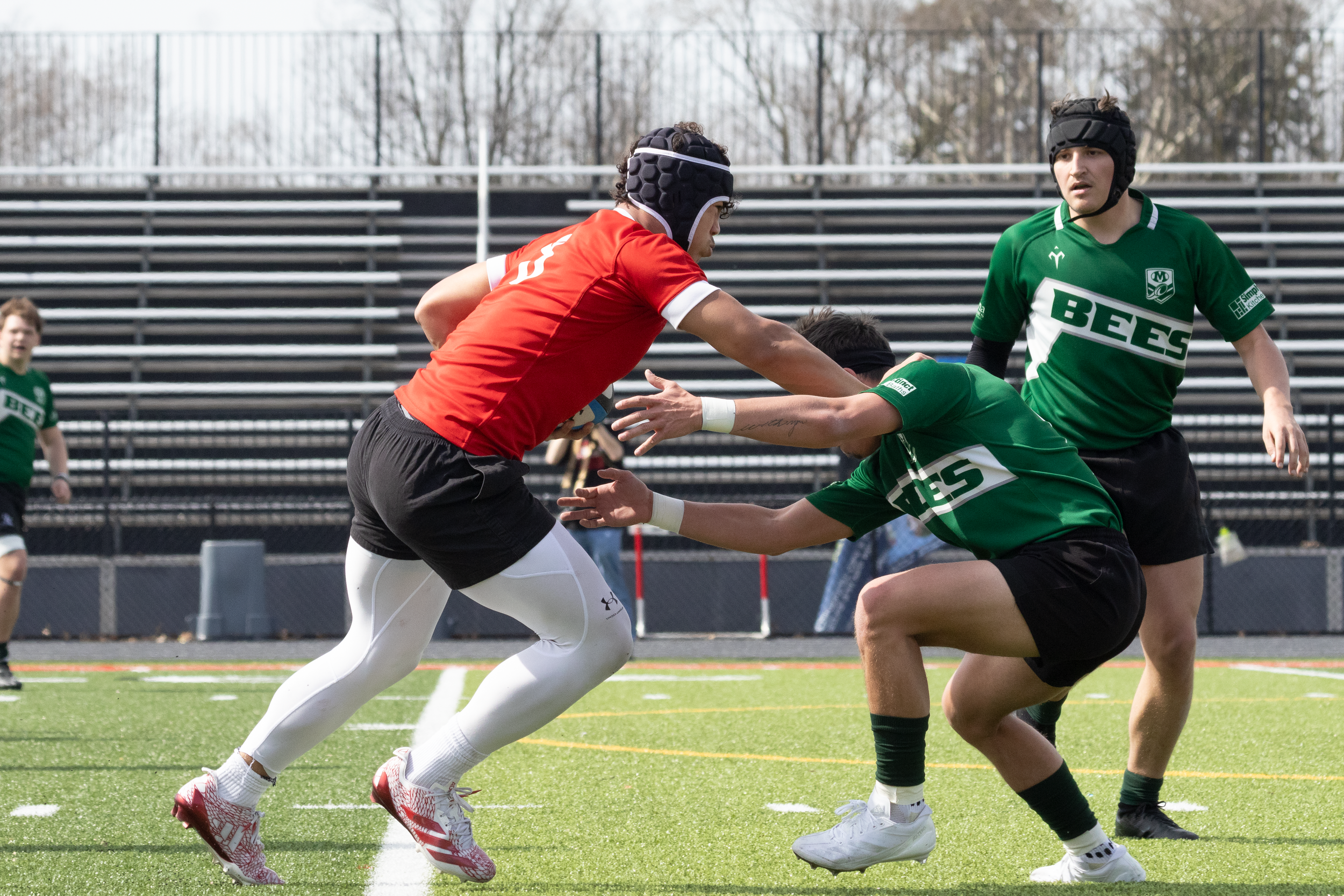 High-intensity rugby match action. Red jersey player evades a tackle from green Bees jerseys on an athletic field.