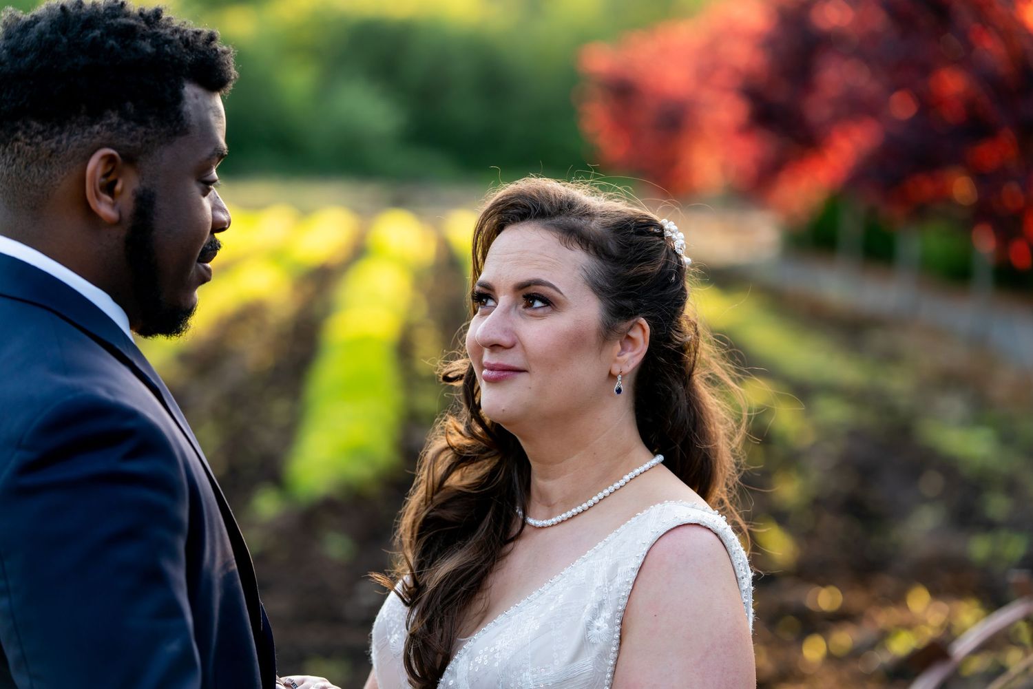 Bride in white dress gazes lovingly at groom in blue suit during outdoor vineyard wedding ceremony.