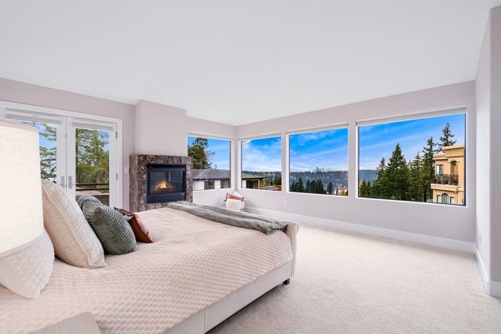 Bright master bedroom with plush carpet, stone fireplace, and large windows offering panoramic views of trees and water.