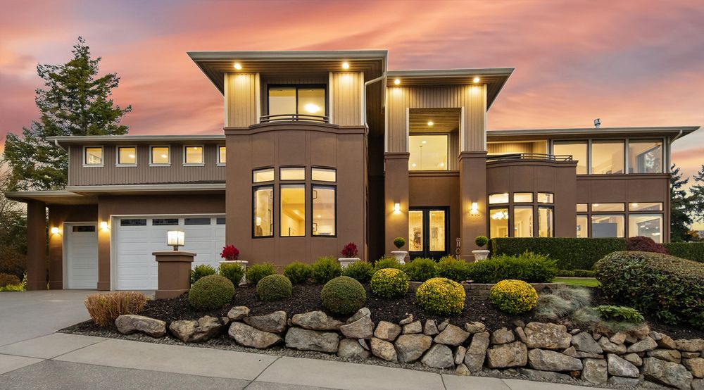 Modern two-story Prairie-style home with stone landscaping, warm interior lighting, and dramatic sunset sky.