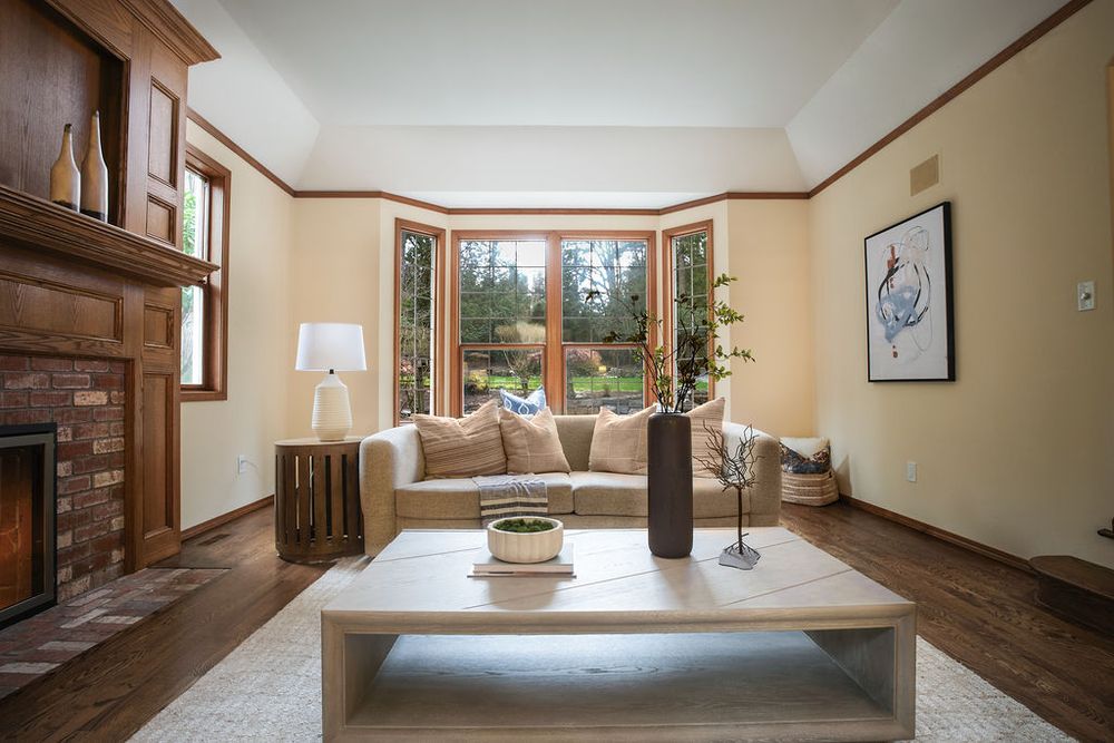 Bright living room with bay windows, brick fireplace, hardwood floors, beige sofa, and wooden coffee table.