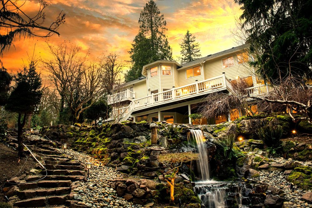 Stunning estate home with waterfall, rock steps, lush greenery, and dramatic sunset sky.