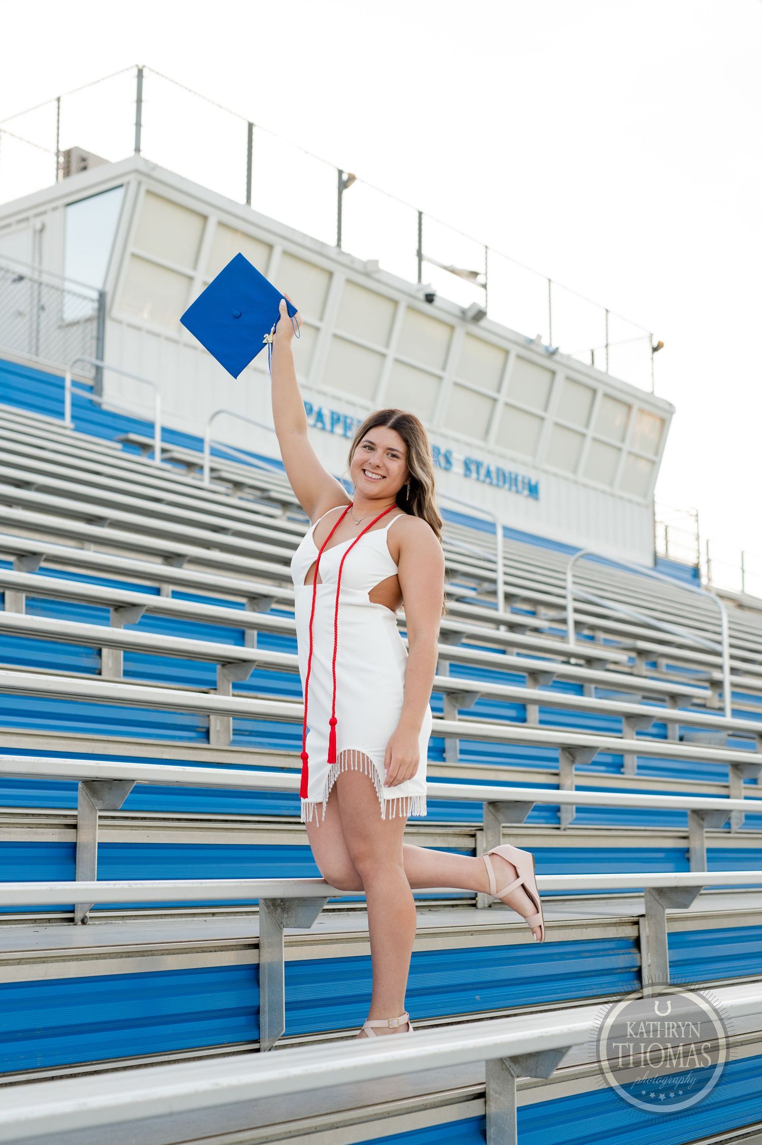 Graduate holds cap high while standing on stadium bleachers in white dress and red stole.