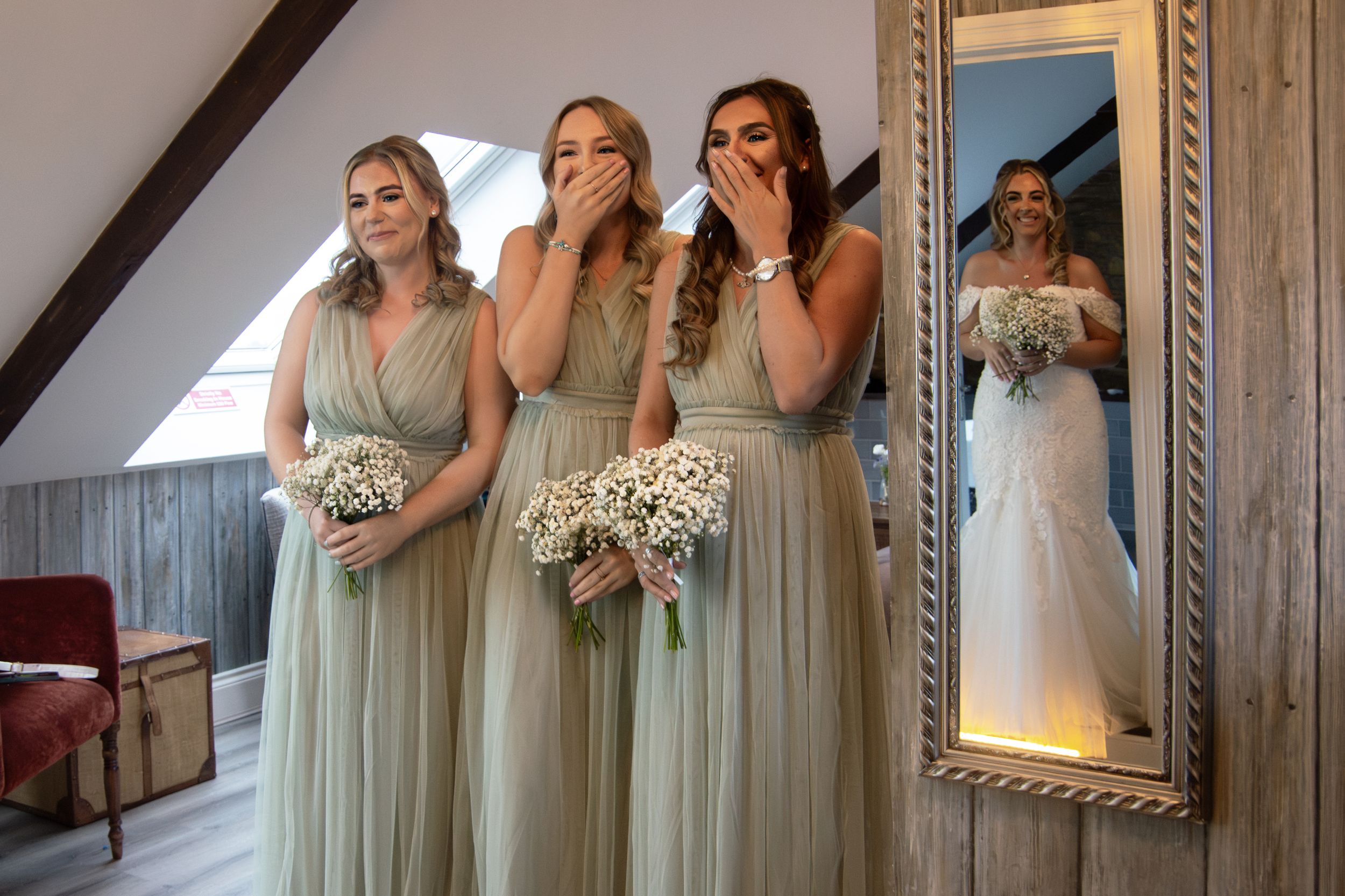 Bridesmaids in sage green dresses share emotional moment while bride is reflected in ornate mirror.