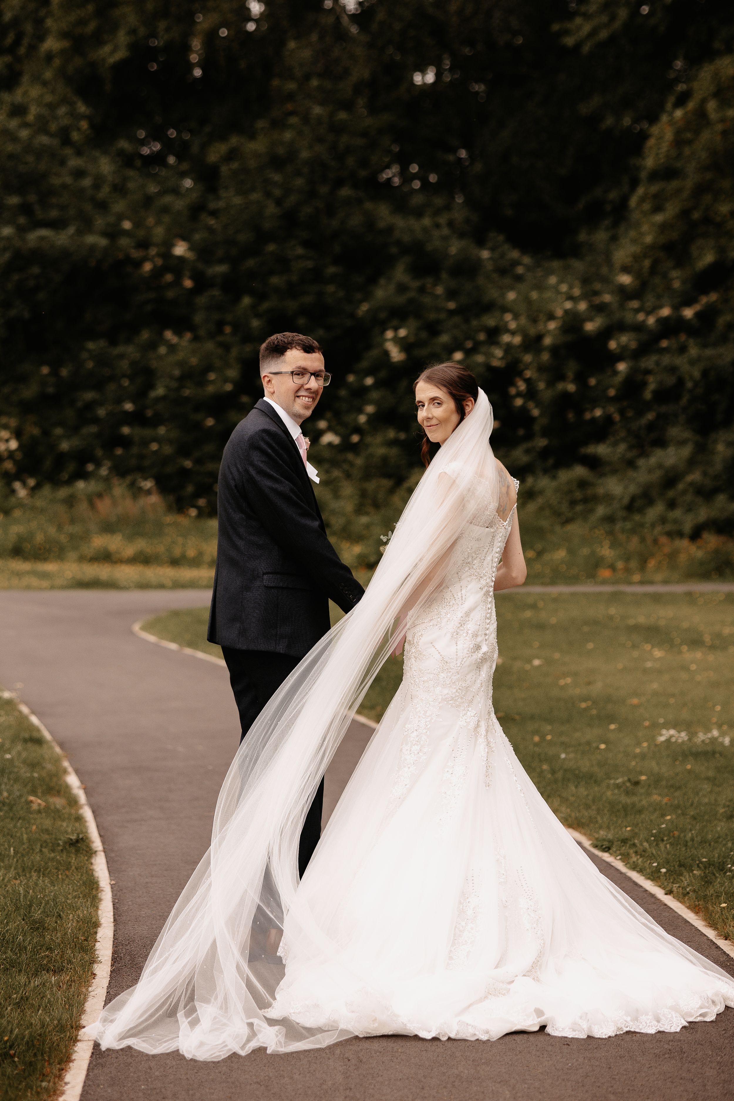 A newly married couple stands on a curved pathway surrounded by lush greenery with a flowing white wedding dress and veil.