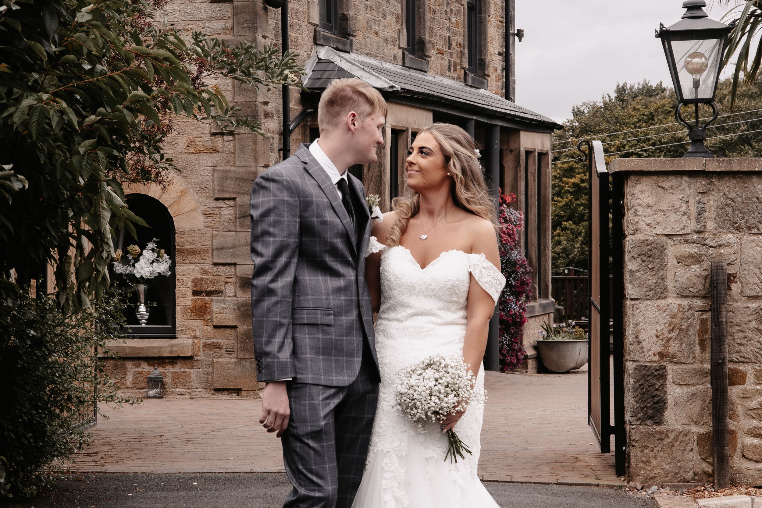 A bride in a strapless white gown and groom in a plaid grey suit stand together outside a stone building.