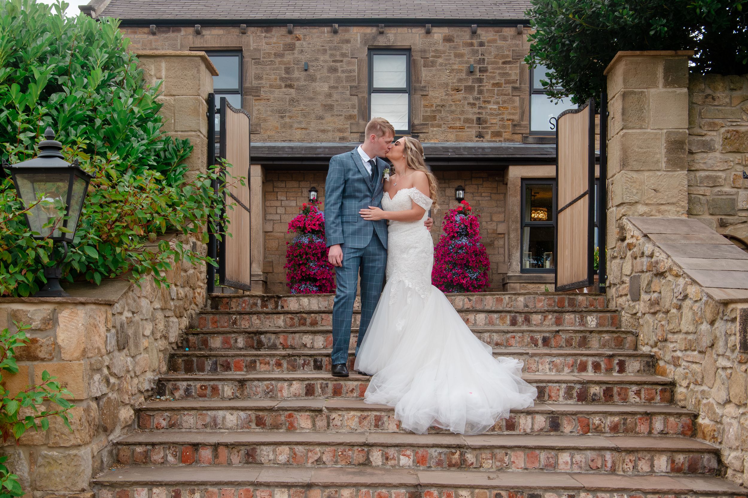 A couple in wedding attire embraces on stone steps of a rustic brick building with red flowers blooming nearby.