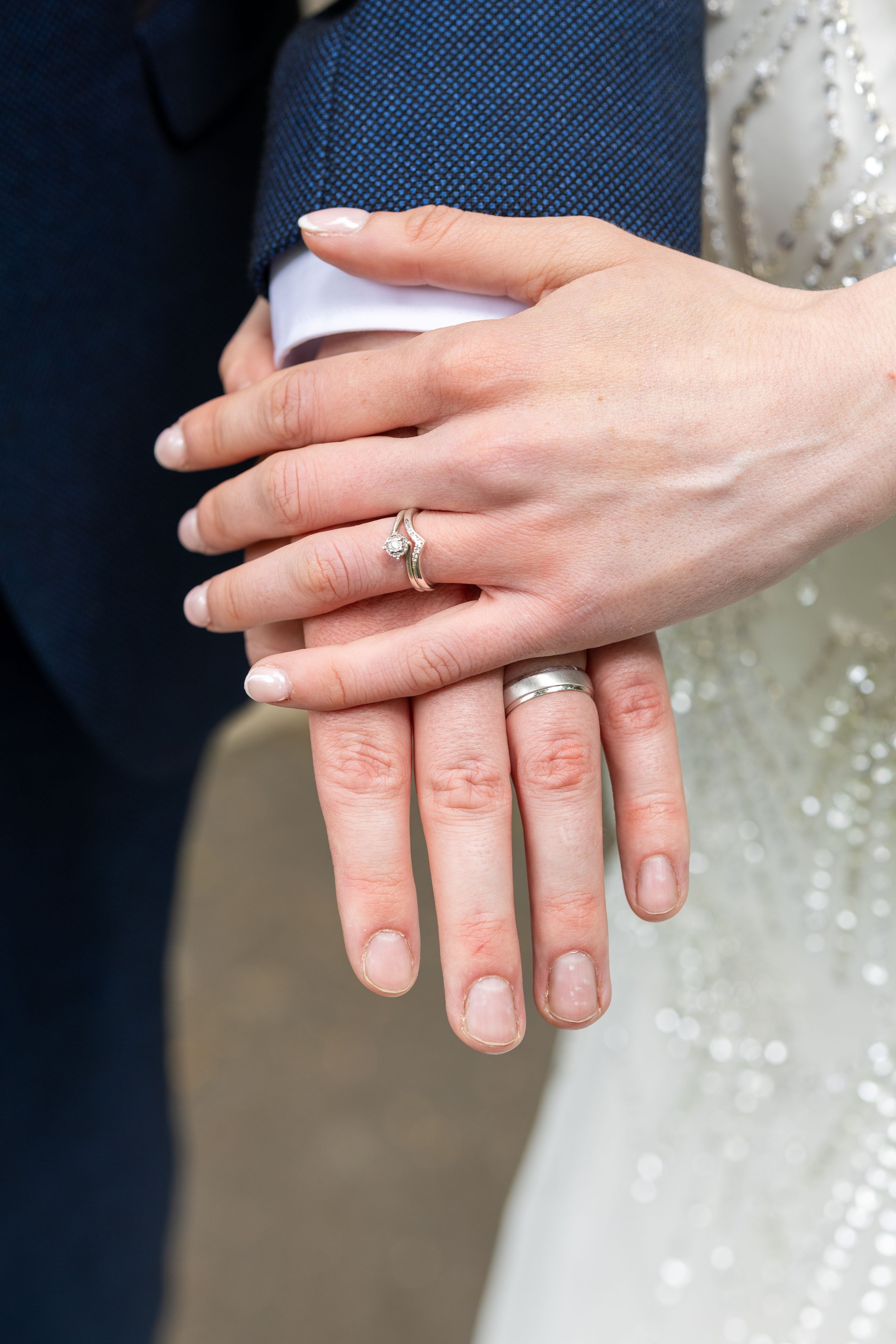 Close-up photo showing hands with wedding rings against a white dress and navy suit.