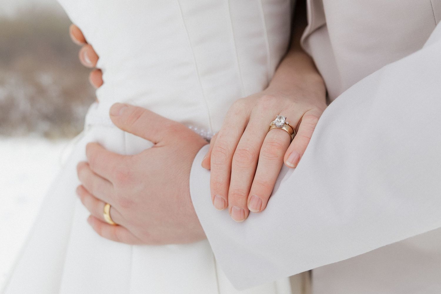 A close-up black and white photo of embracing couple showing hands, with a white dress and small flowers visible.