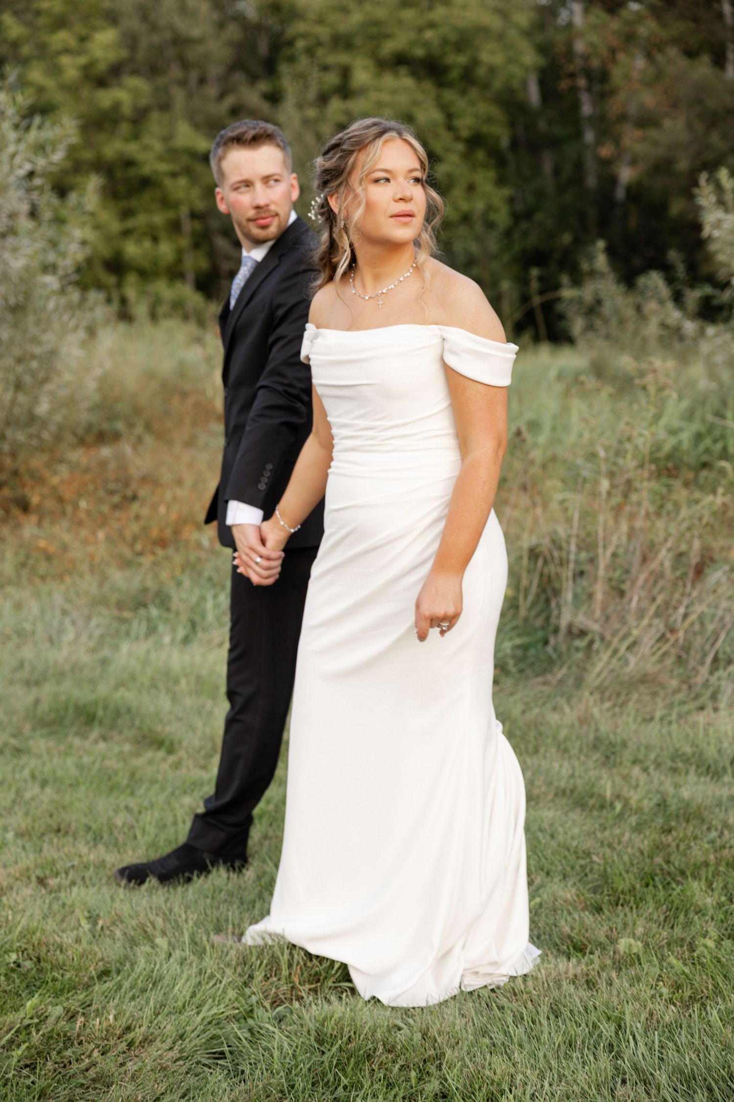 Romantic wedding photography in St.Paul, MN featuring a bride in an off-the-shoulder gown and a groom in a black suit walking in a field.