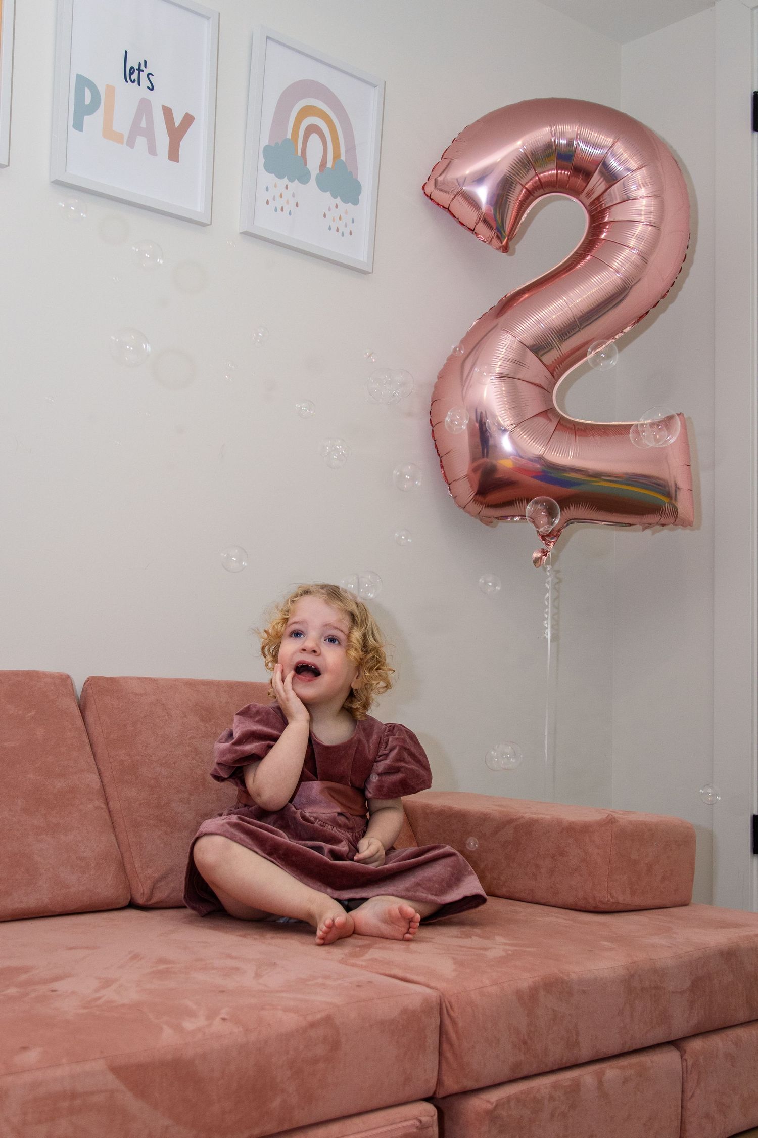 Rose gold number 2 balloon decorates a room with rainbow wall art and a child sitting excitedly on pink couch.