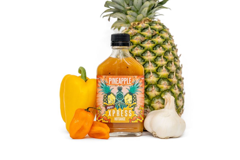 A bottle of hot sauce arranged with fresh pineapple, yellow bell pepper, and garlic on a white background.