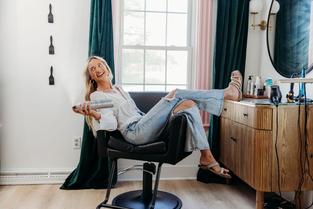 Casual figure lounges sideways in black styling chair wearing light denim and white top near emerald curtains.