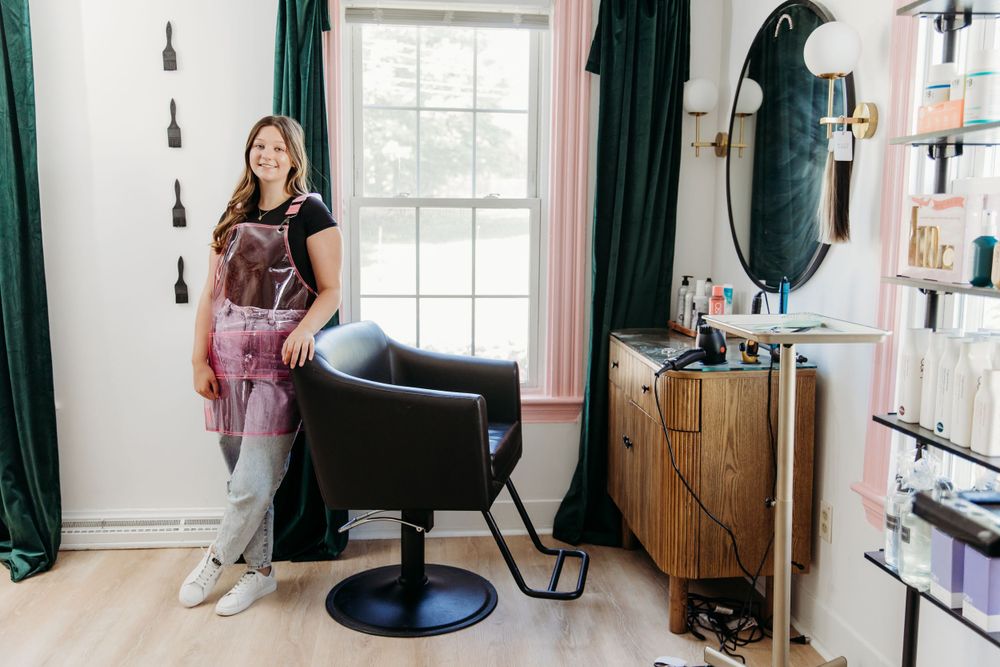 A stylist stands in a cozy salon space with a black styling chair, dark curtains, and wooden storage cabinet.