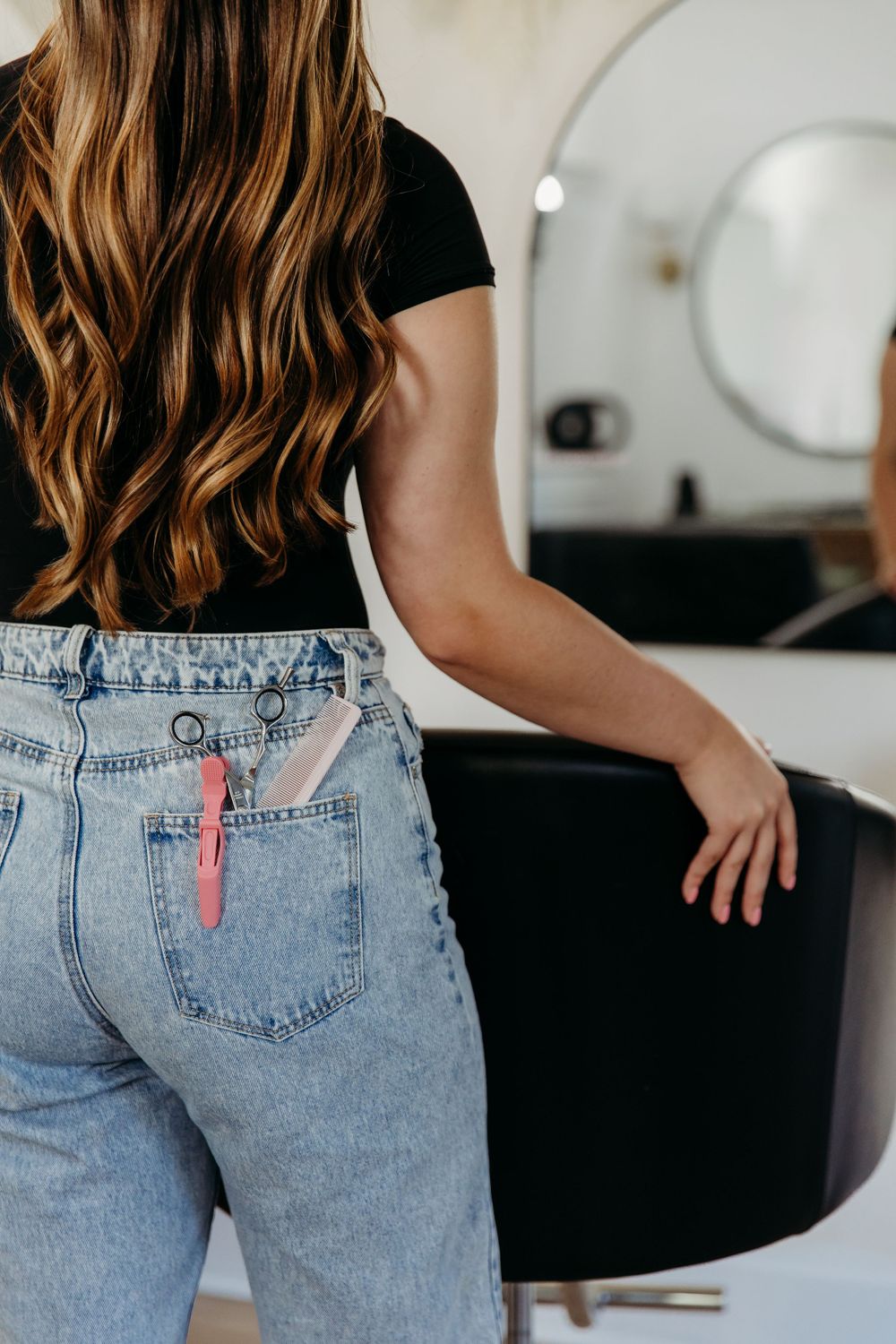 Hair stylist in black shirt and light wash jeans stands next to salon chair with styling tools in back pocket.