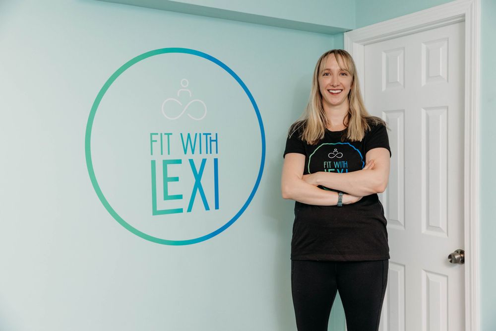 Fitness instructor wearing black workout clothes poses with crossed arms in front of turquoise wall with circle logo.