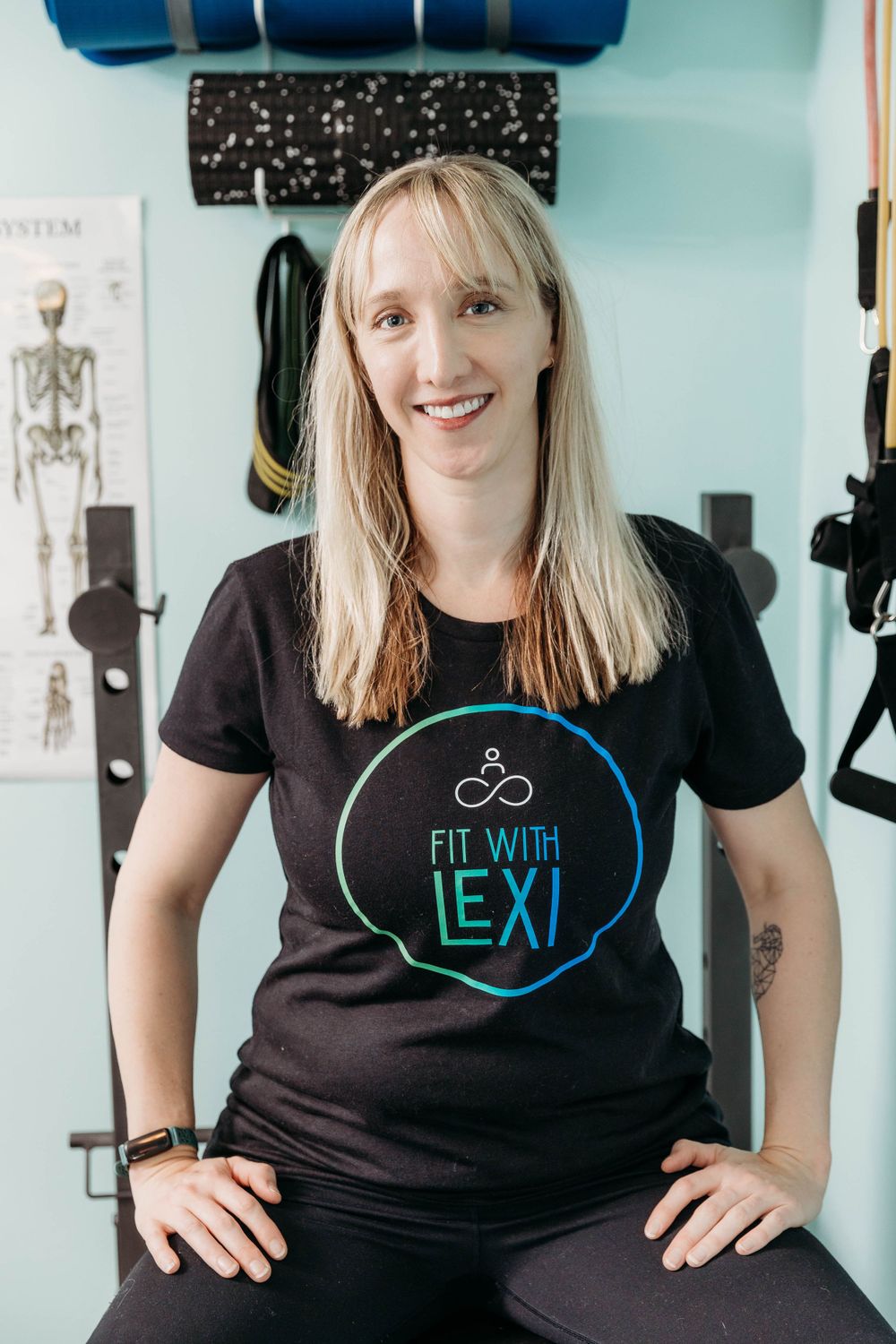 A fitness instructor wearing a black t-shirt with 'Fit with Lexi' logo sits in an exercise studio setting.