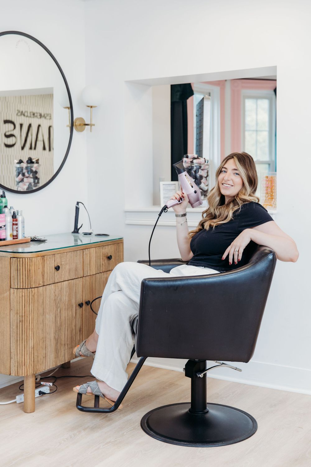 A bright modern salon interior with a black styling chair and wooden cabinet beside a decorative wall mirror.