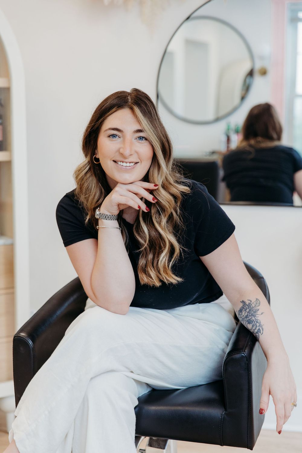 Woman with long wavy hair wearing black t-shirt and white pants sits casually in leather salon chair.