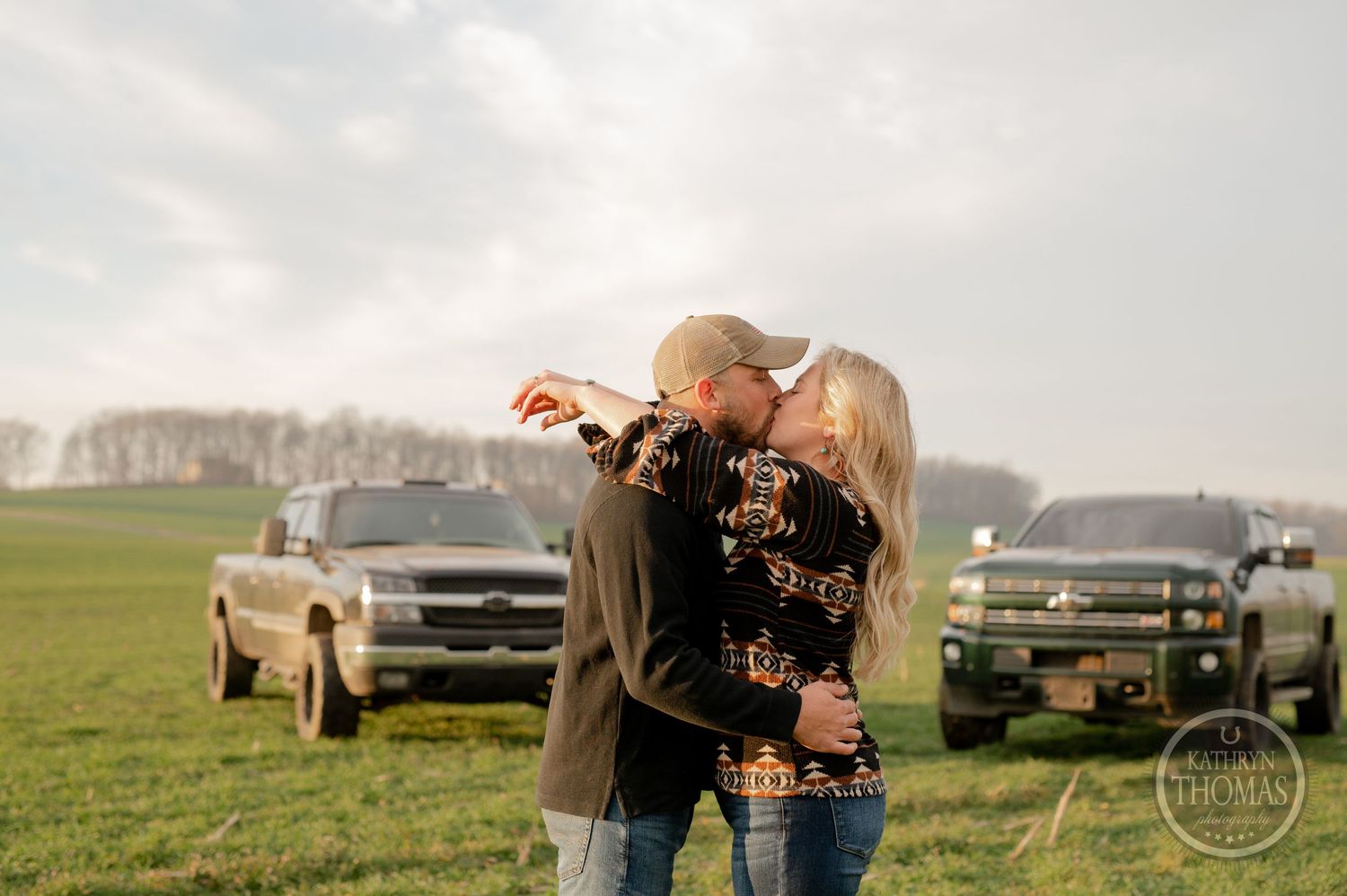 A couple shares a romantic moment in a field at sunset with parked trucks behind them.