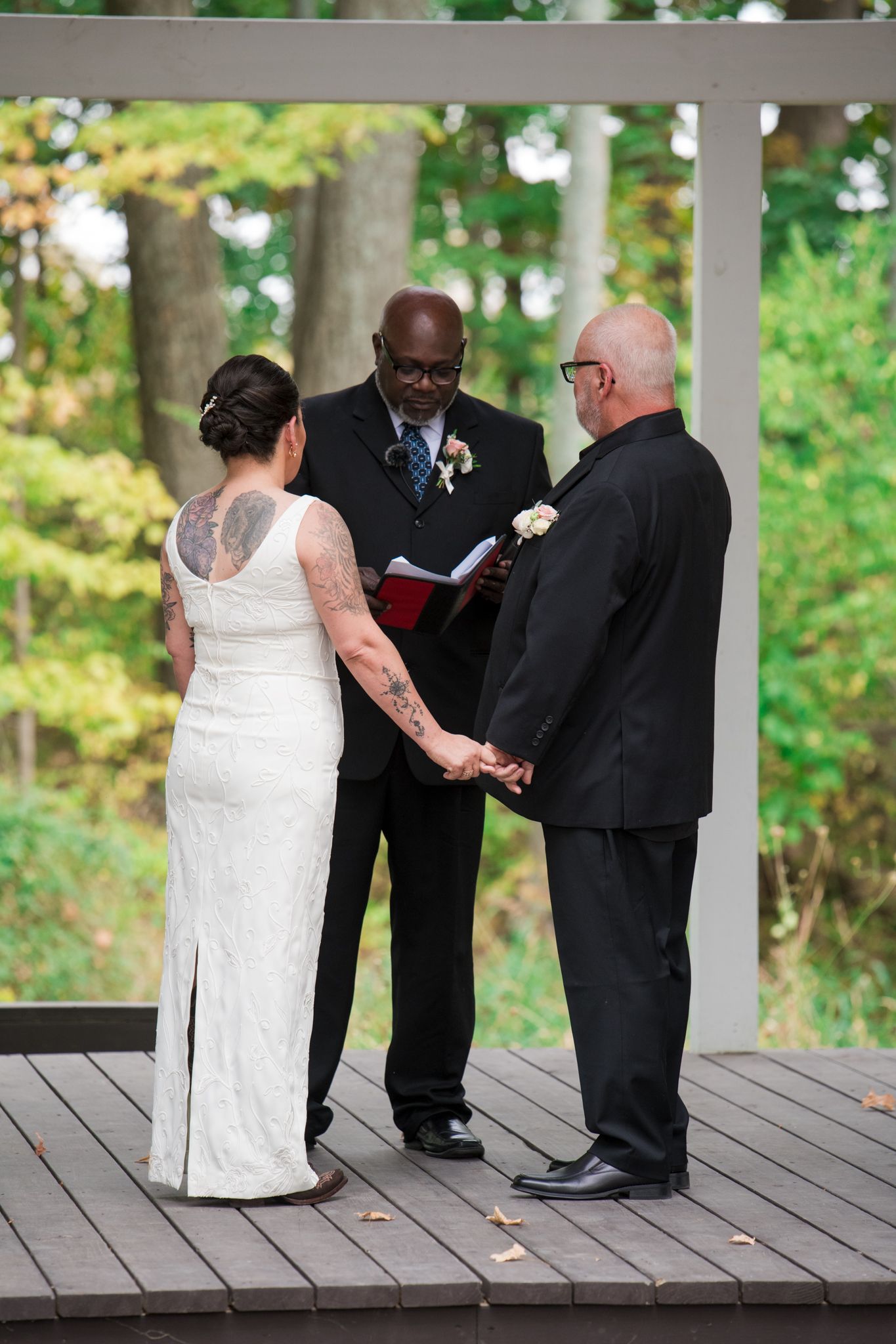 A bride in a flowing white gown walks along a tree-lined path with companion.