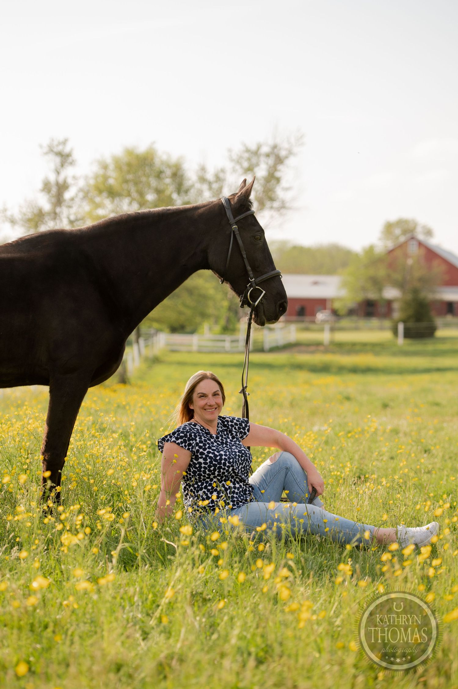 A beautiful black horse stands beside someone relaxing in a field of yellow wildflowers.