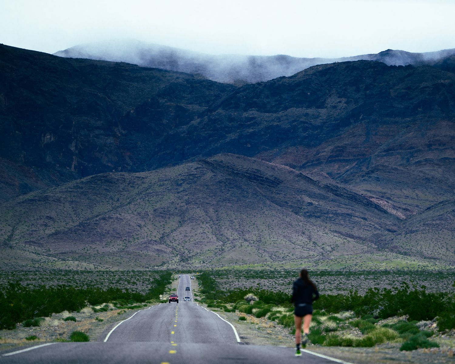 Long empty road stretches toward misty mountains in the Mojave Desert with a lone cyclist in the distance.