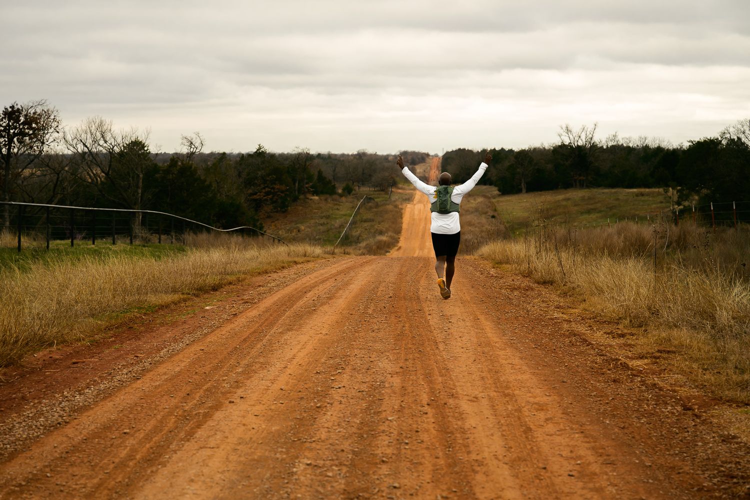 A lone runner with raised arms walks on a dusty dirt road through rural countryside on an overcast day during the Mid South Gravel 50k race.