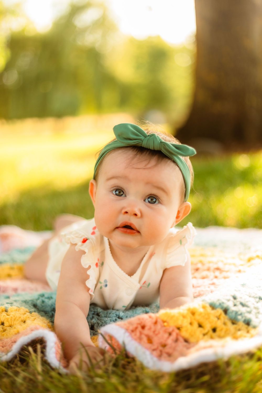 Adorable baby girl in green bow headband lying on colorful crochet blanket outdoors in sunny park.