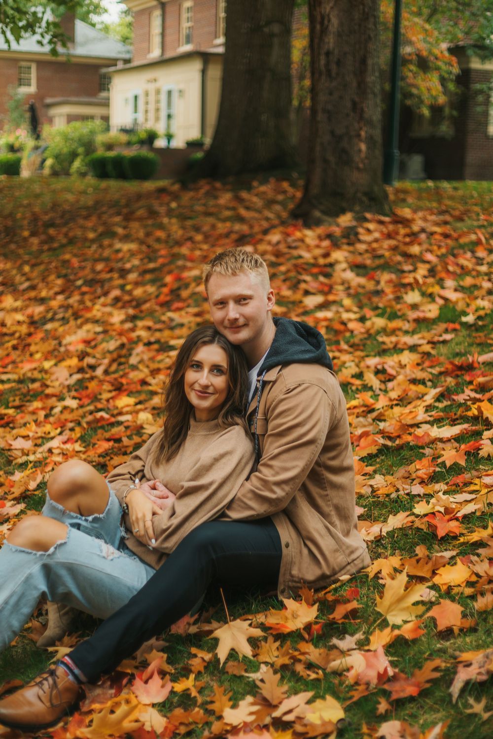Couple sitting together on grass covered in colorful autumn leaves, embracing warmly in front of a brick building.